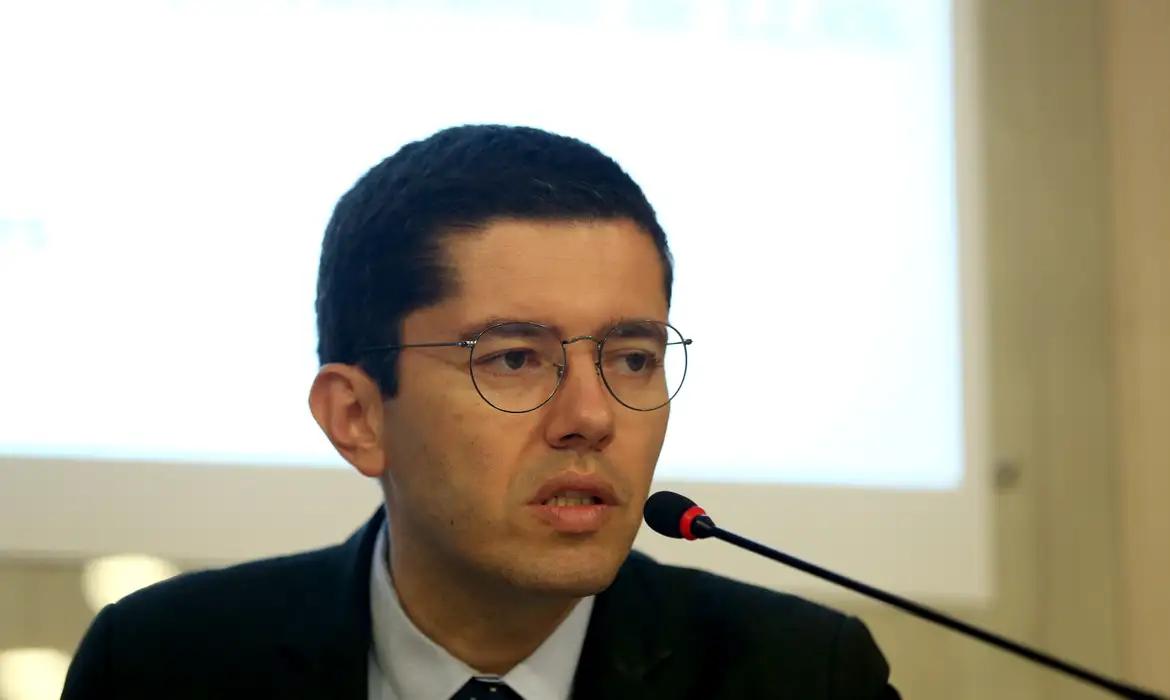 Homem de terno e óculos fala ao microfone durante evento ou coletiva em ambiente interno.
